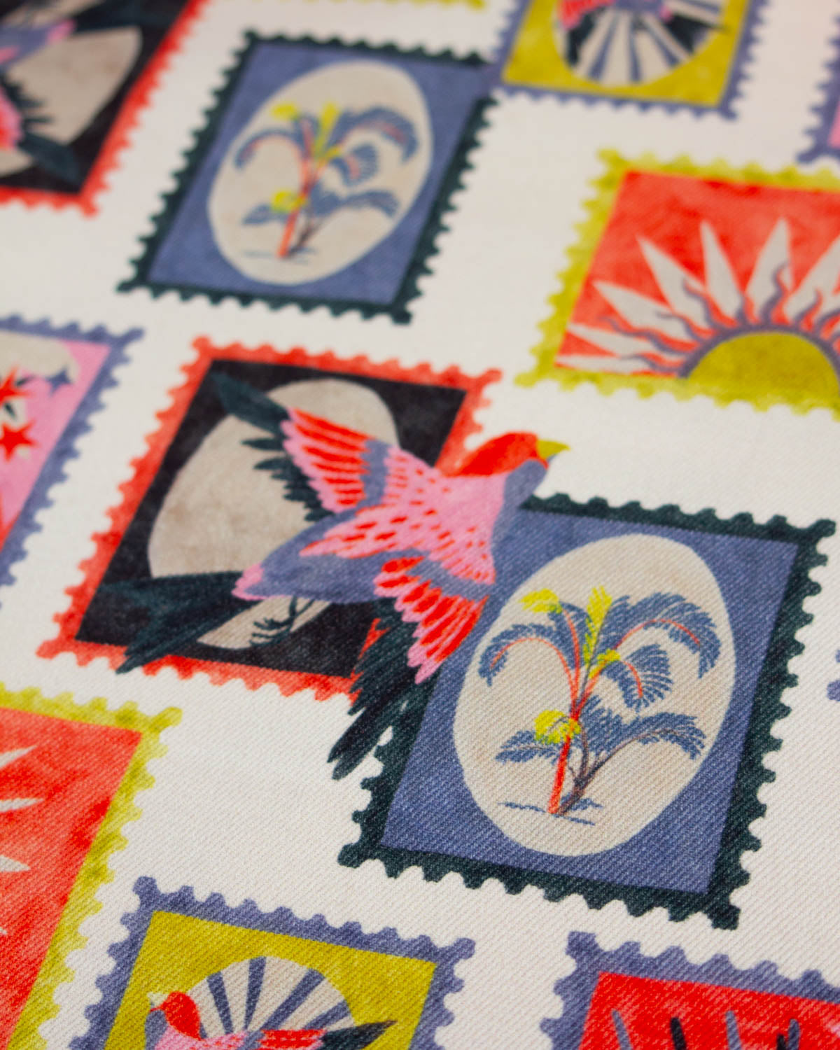 A close-up of a cotton denim fabric print featuring colorful postage-stamp-like squares with tropical motifs, including palm trees, birds, and suns on a white base.