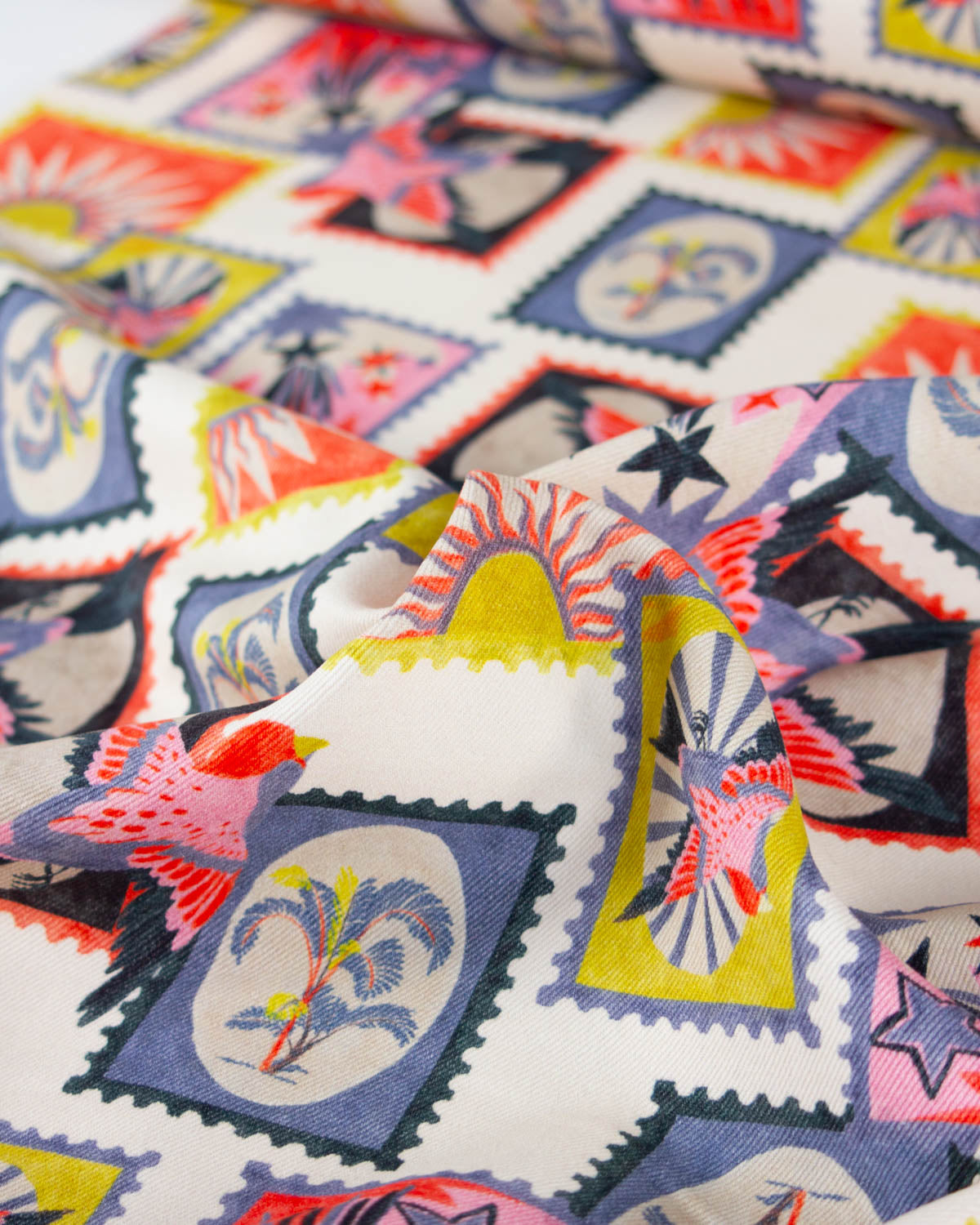 A close-up of a multicolor cotton denim fabric with a vintage postage stamp pattern featuring birds, palm trees, stars, and suns on a cream background.