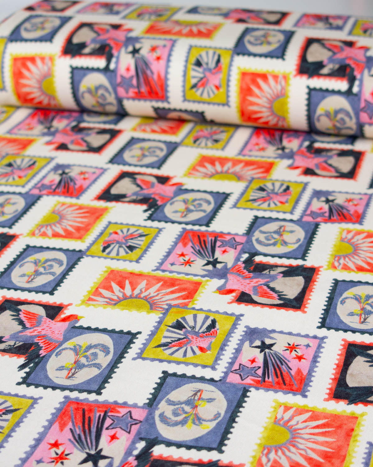A close-up of a patterned cotton denim fabric featuring a repeating design of postage stamps with colourful suns, trees and shooting stars on a white background.