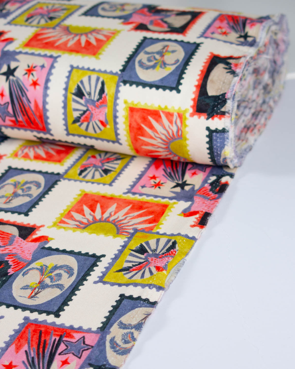 A roll of cotton denim fabric with a colorful print of postage stamps, featuring various designs in red, yellow, blue, pink, and black on an off-white background.