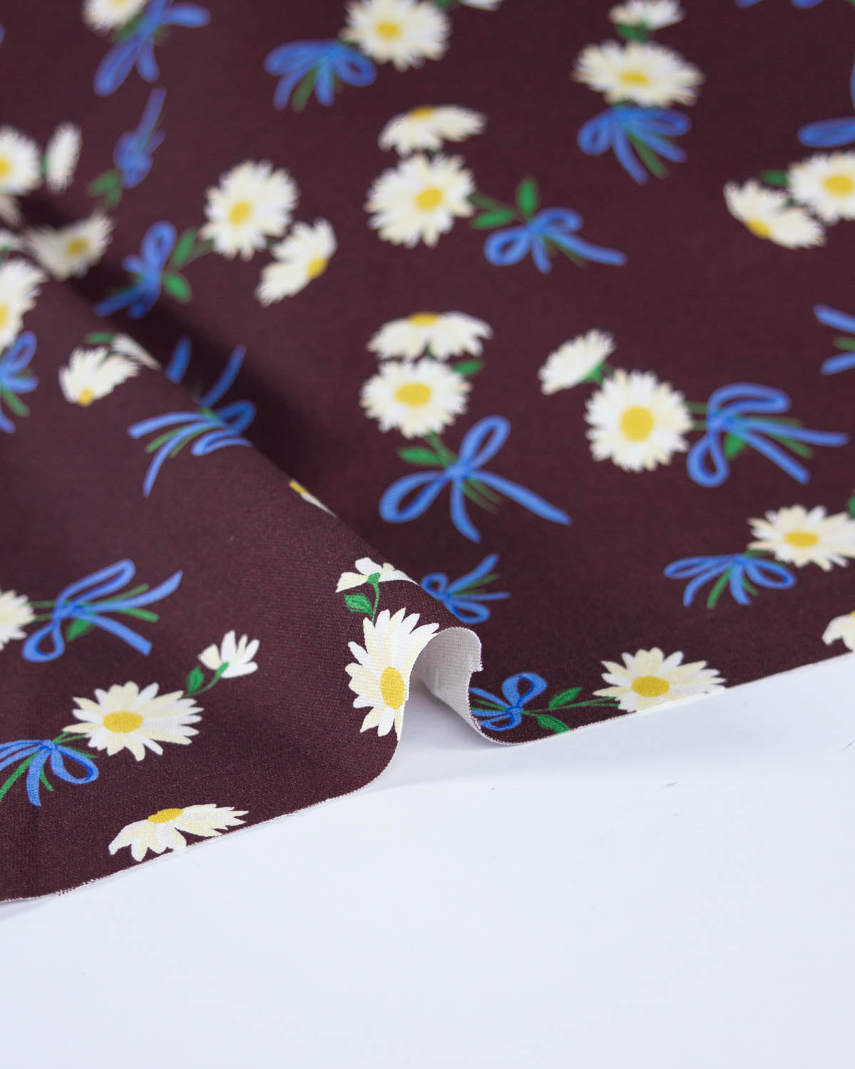 A close-up of a dark brown cotton canvas fabric with a repeating pattern of white daisies, blue bows, and green leaves, draped over a white surface.