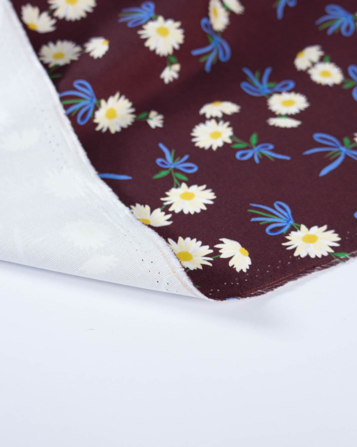 A close-up of a folded cotton canvas fabric, showing a dark burgundy/maroon side with a white, yellow, and blue daisy floral pattern, and a white reverse side with subtle tonal floral embroidery.