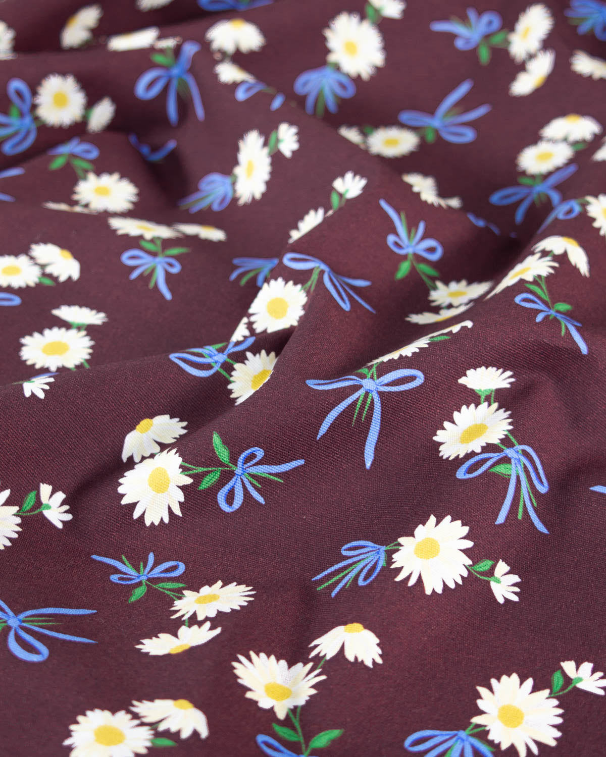 A close-up of a draped cotton canvas fabric with a dark brown base, featuring a repeating pattern of white daisies with yellow centers, blue ribbon bows, and small green leaves.