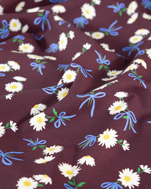 A close-up of a draped cotton canvas fabric with a dark brown base, featuring a repeating pattern of white daisies with yellow centers, blue ribbon bows, and small green leaves.