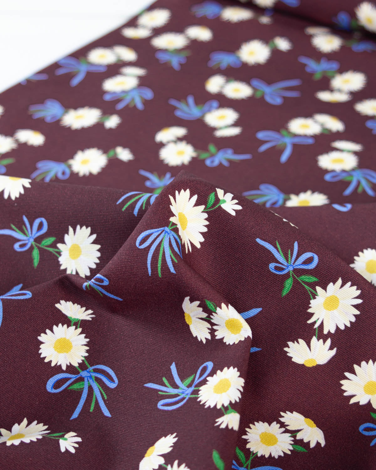 A close-up of a dark brown cotton canvas fabric with a repeating pattern of white daisies, blue bows, and green leaves.