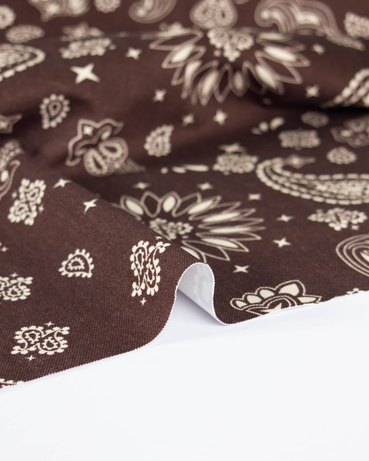 A close-up of a brown cotton denim fabric with a cream paisley and floral print, folded over to show its texture and white reverse side against a white background.