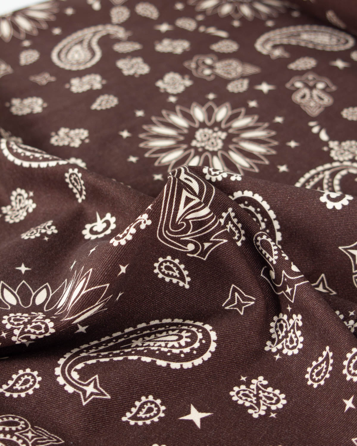 A close-up of a brown cotton denim fabric with a white paisley and floral bandana print, showing the material's texture and pattern details.