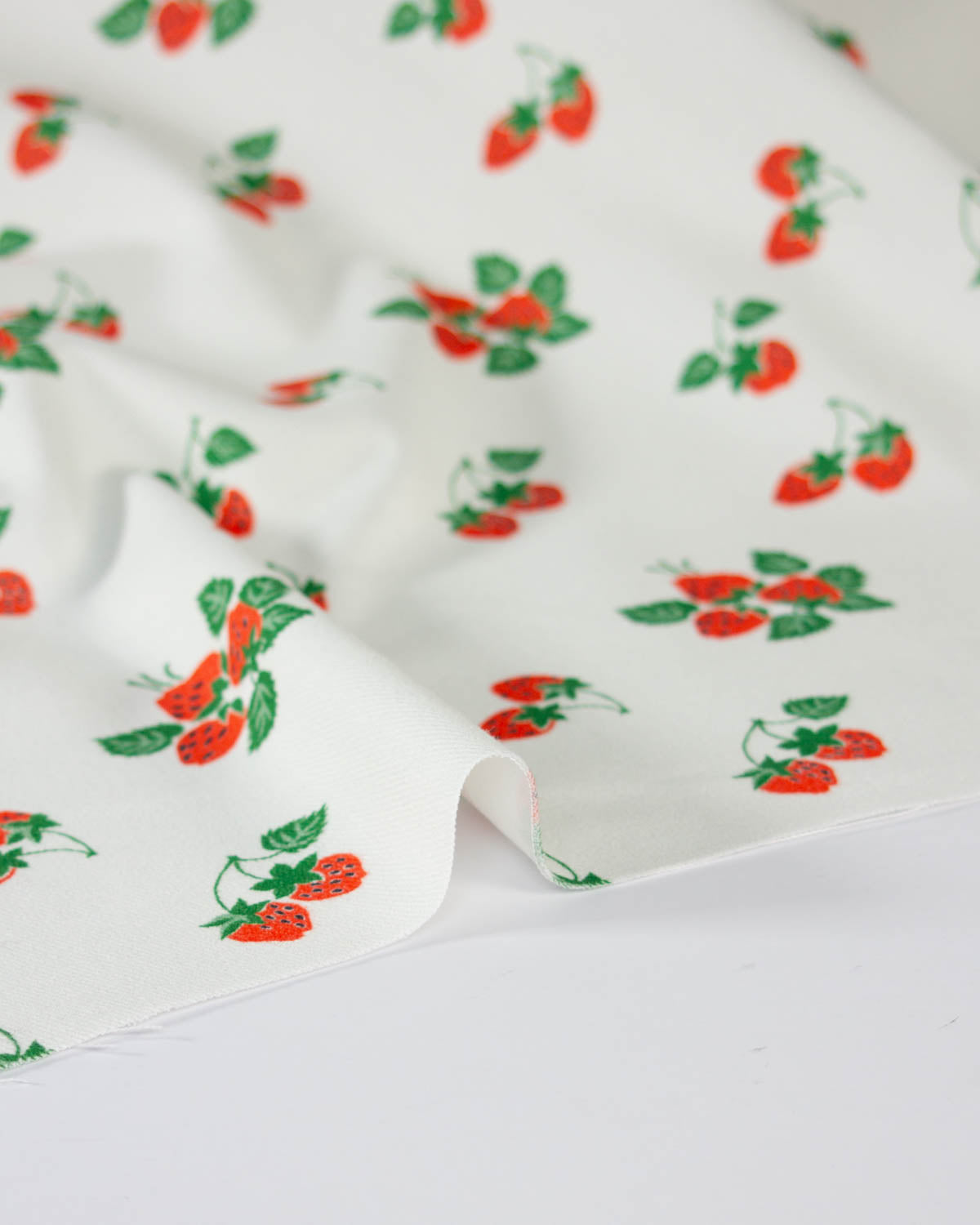 White cotton denim fabric with a repeating pattern of red strawberries and green leaves, draped to show its texture and print.