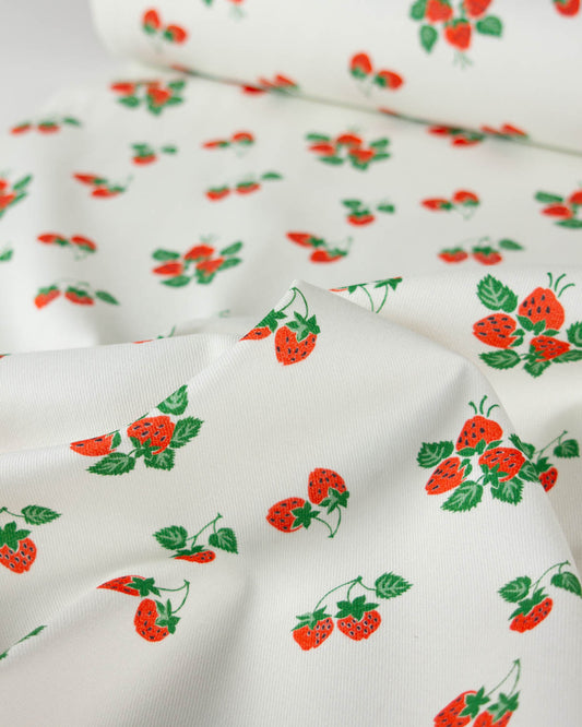 White cotton denim fabric with a repeating pattern of red strawberries and green leaves, shown folded with a rolled section in the background.
