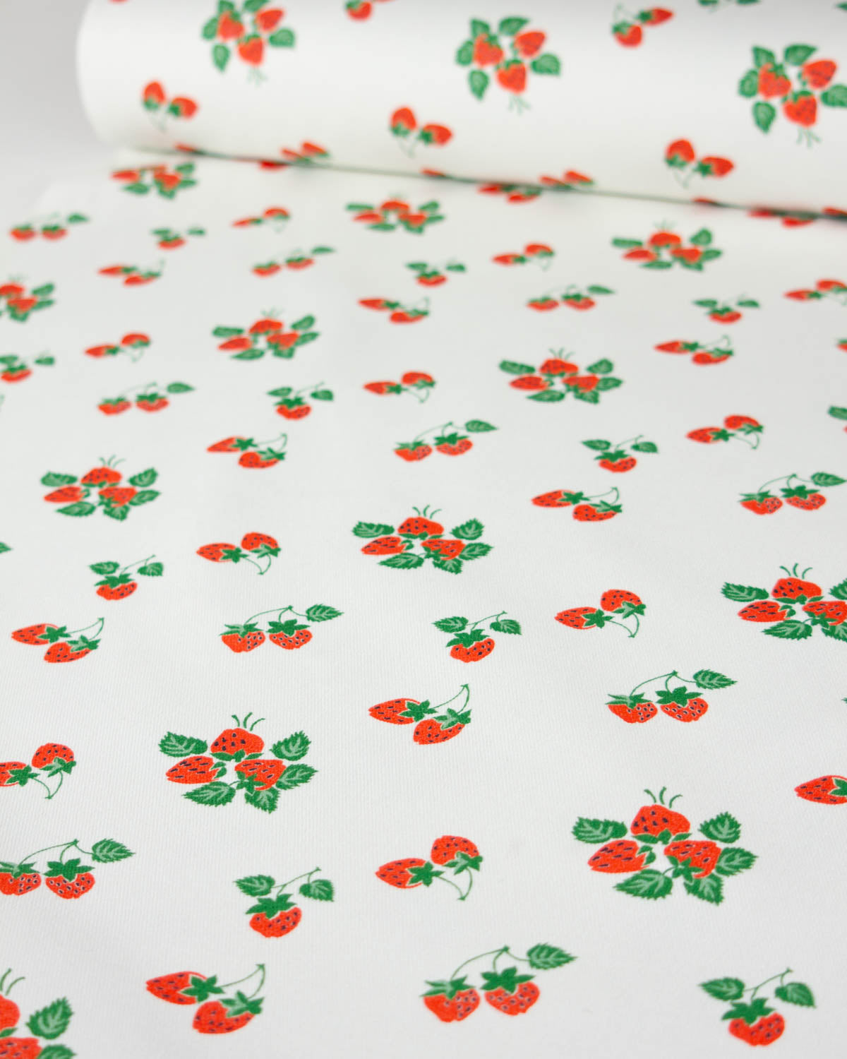 White cotton denim fabric with a repeating pattern of red strawberries and green leaves, shown with one roll partially unrolled at the top.