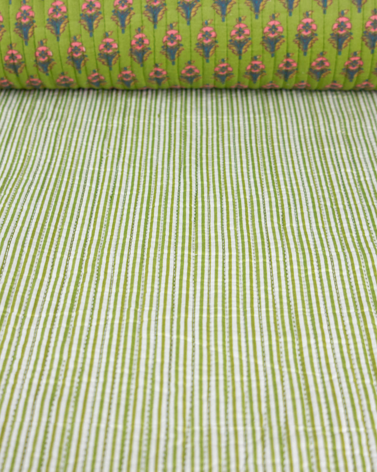 A close-up of a green patterned quilted. cotton fabric, showing one side with fine vertical white and green stripes and a second side with a repeating floral motif in pink, orange, and green.