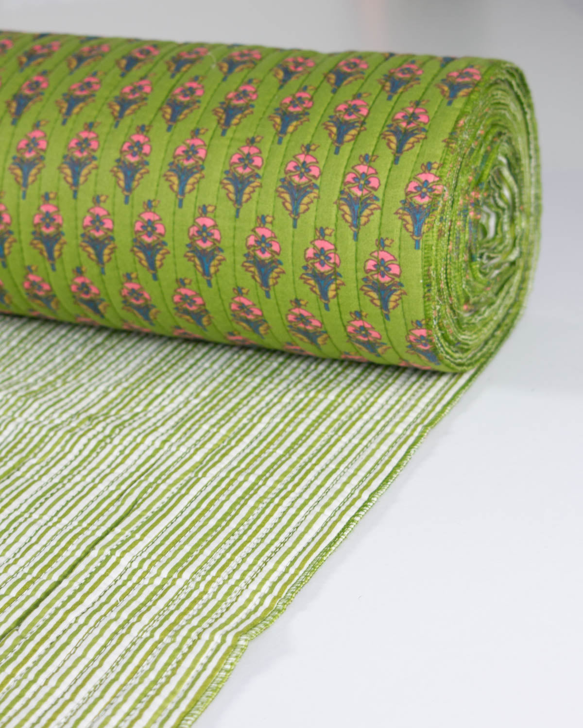 A roll of green quilted cotton fabric with a repeating pink and blue floral pattern, shown partially unrolled on a white surface.