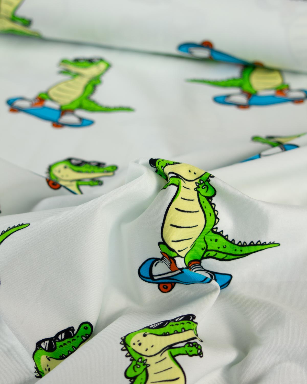 White cotton jersey fabric printed with a playful pattern of cartoon alligators riding blue skateboards, shown in a close-up draped view.