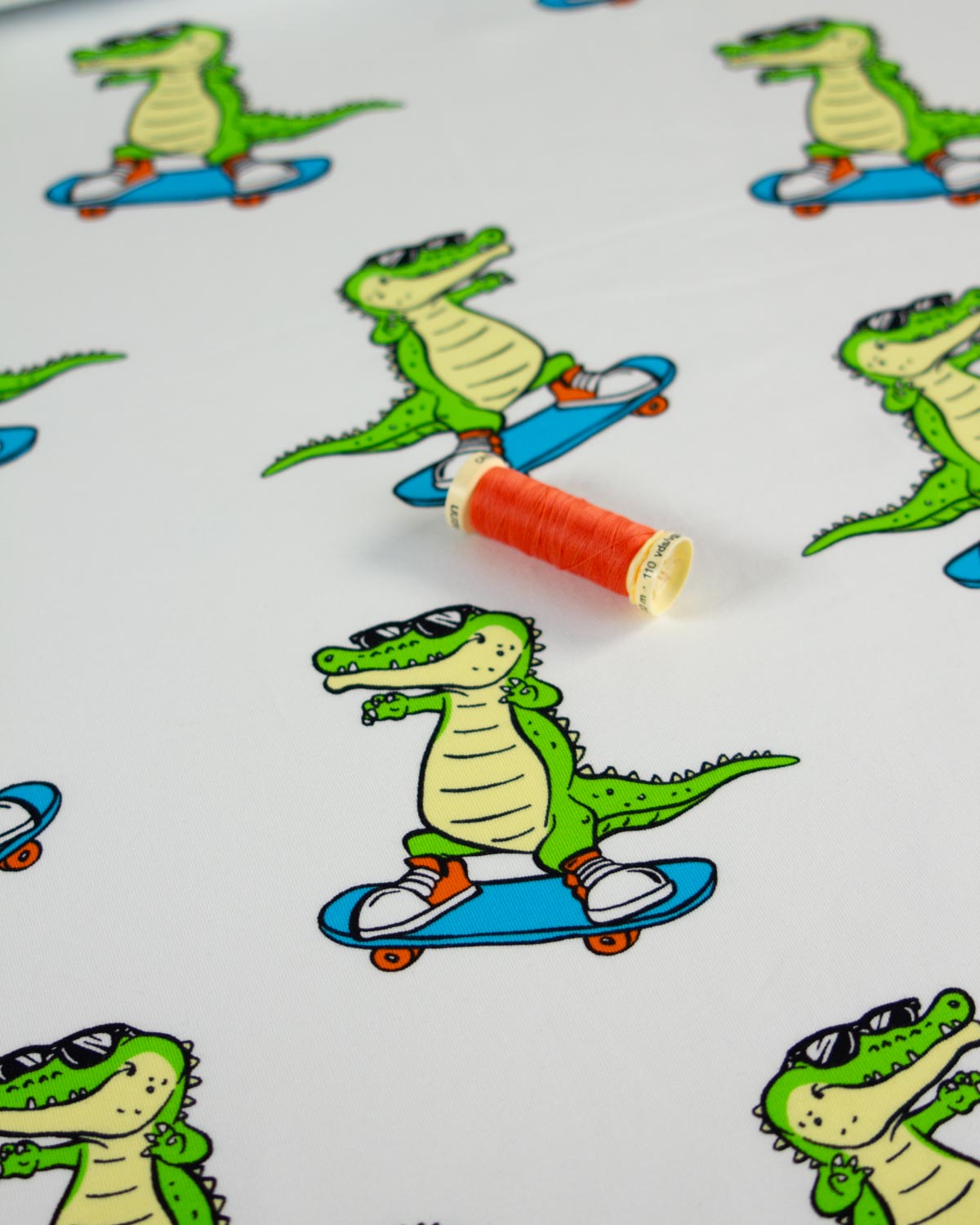 A close-up of a white cotton jersey fabric surface printed with a repeating cartoon alligator riding a blue skateboard, with a spool of orange thread placed on top.