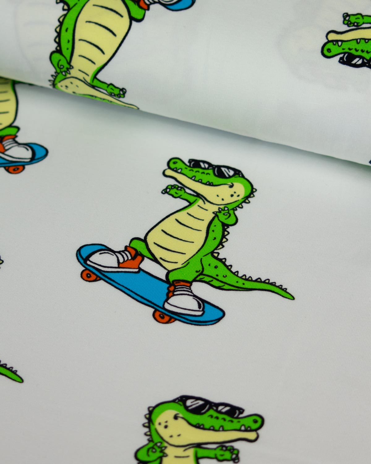 White fabric with a repeating cartoon alligator/crocodile graphic riding a blue skateboard, shown in a close-up roll.