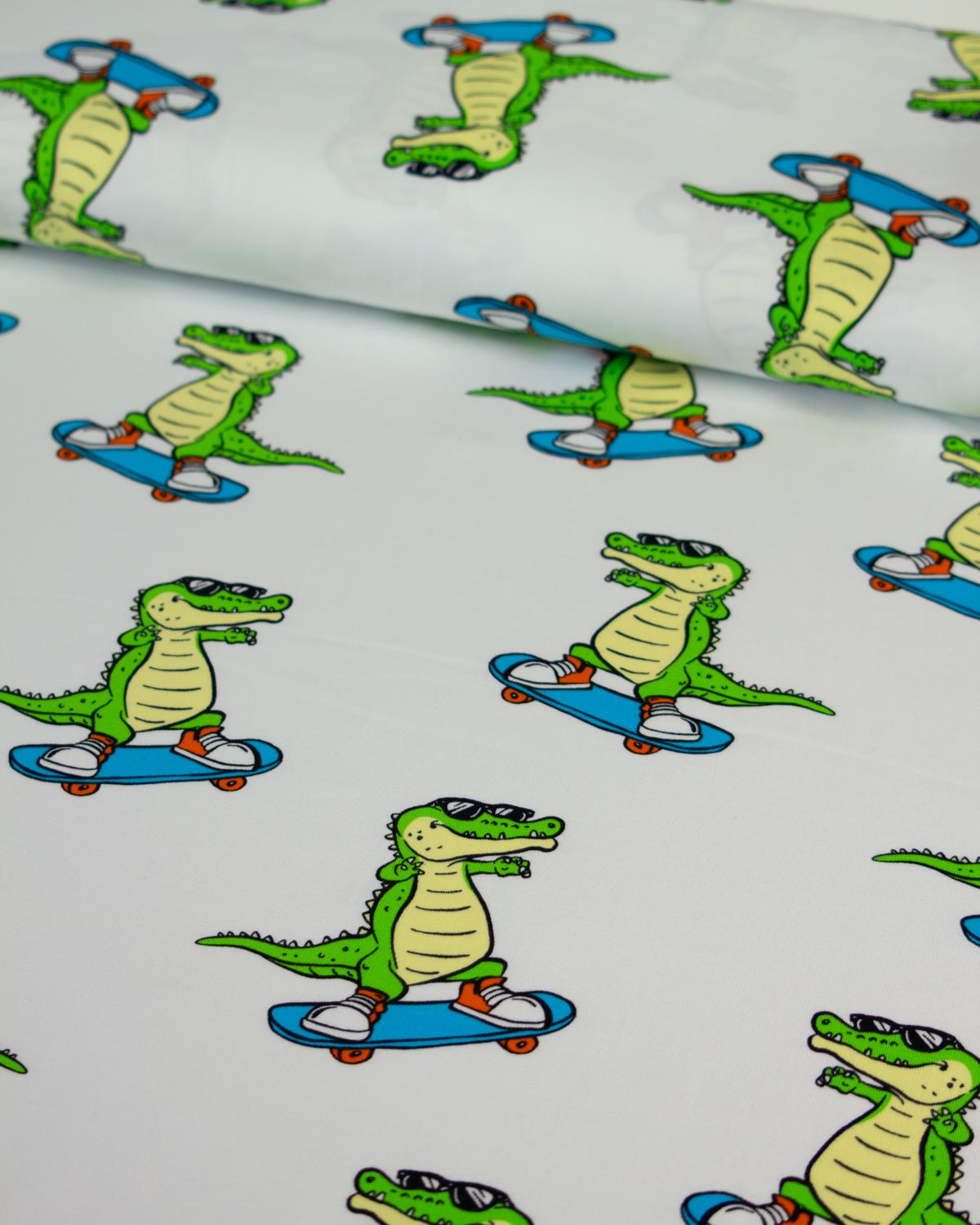 White cotton jersey fabric with a repeating cartoon alligator/crocodile riding a blue skateboard pattern, shown in a close-up flat view with a rolled section at the top.