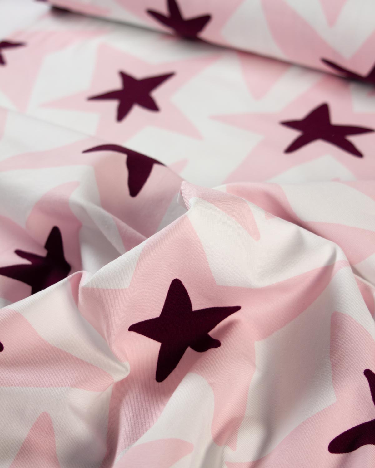 A close-up of a cotton jersey fabric surface with a repeating star pattern in dark plum on a soft pink and white background.