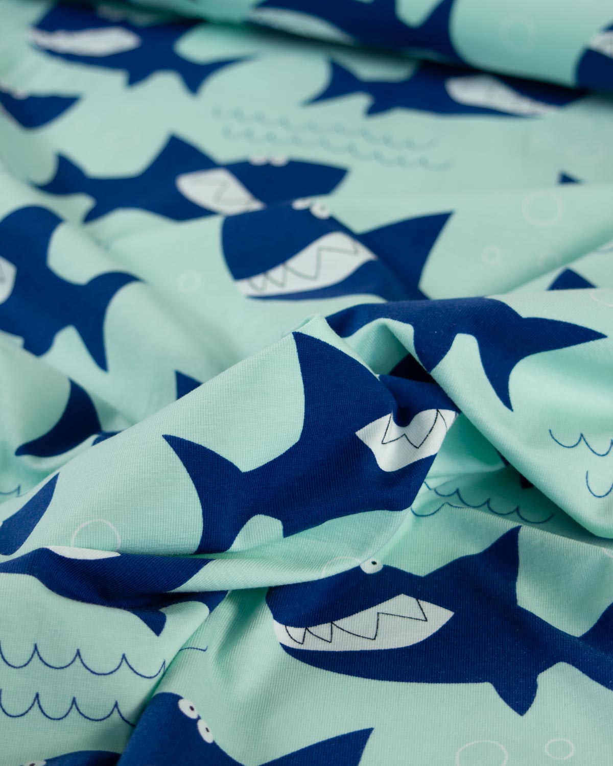 A close-up of a draped cotton jersey  fabric featuring a playful pattern of dark blue sharks swimming on a light mint green background with simple white details.
