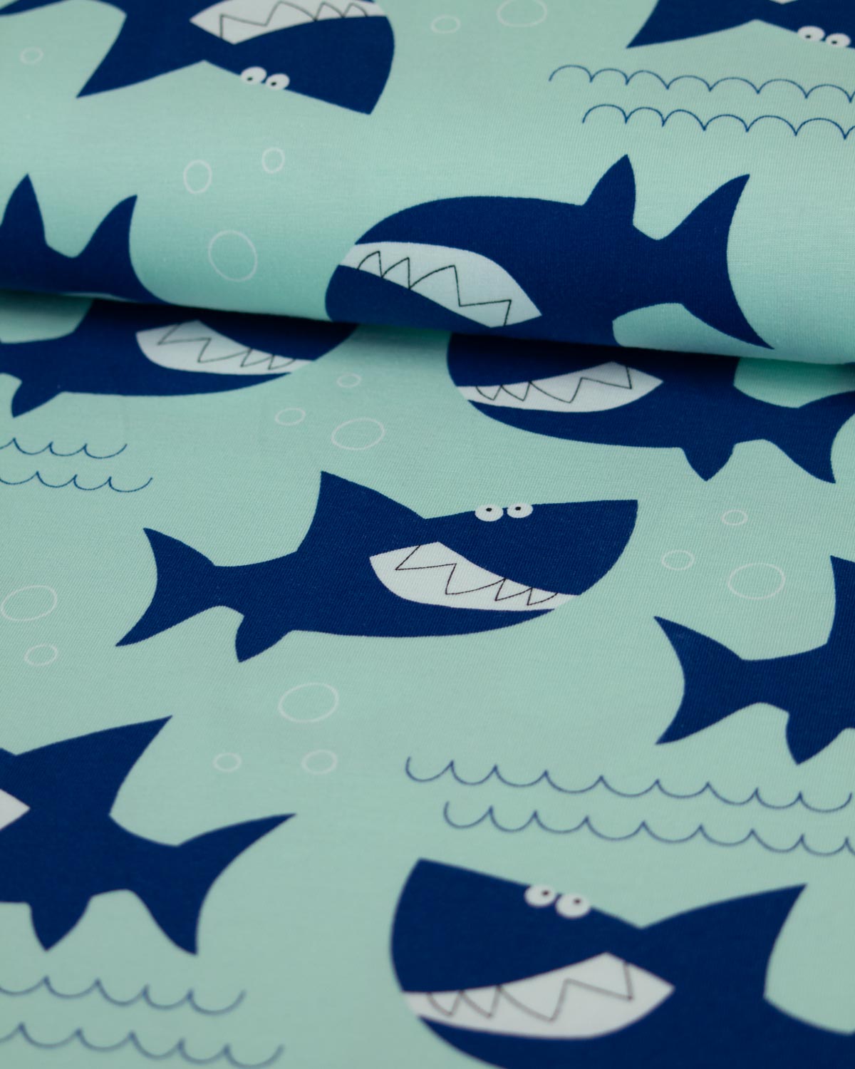 A close-up of a cotton jersey  fabric with a repeating shark print. The background is a light mint green, with dark navy blue cartoon sharks and small white details like teeth, eyes, and bubbles.