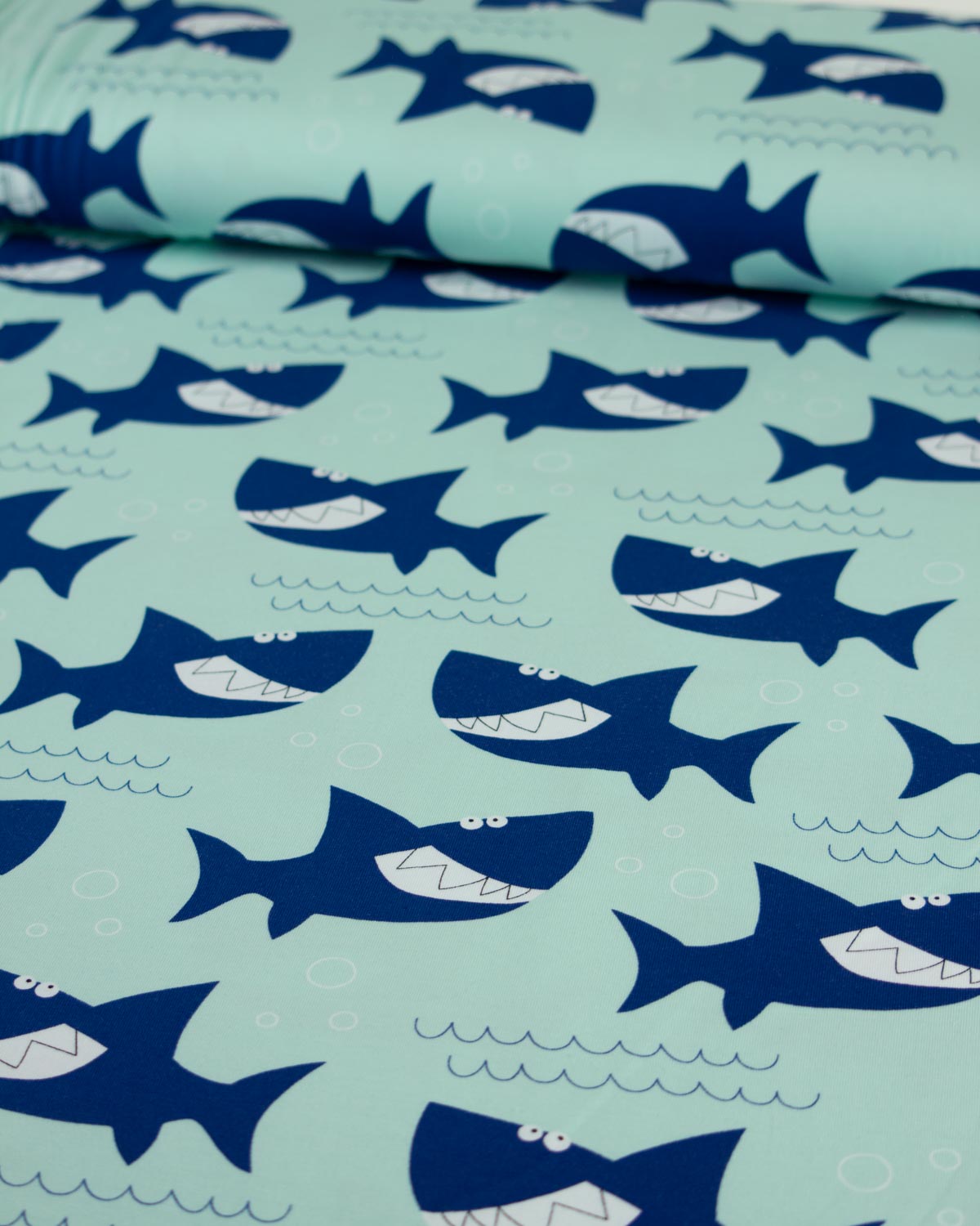 A close-up of a light mint green cotton jersey  fabric printed with a repeating pattern of dark blue cartoon sharks and small white wave and bubble details.