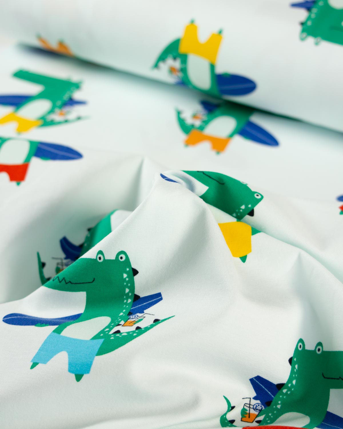 White cotton jersey fabric with a colorful cartoon crocodile print, shown in a close-up with folds and a rolled section in the background.