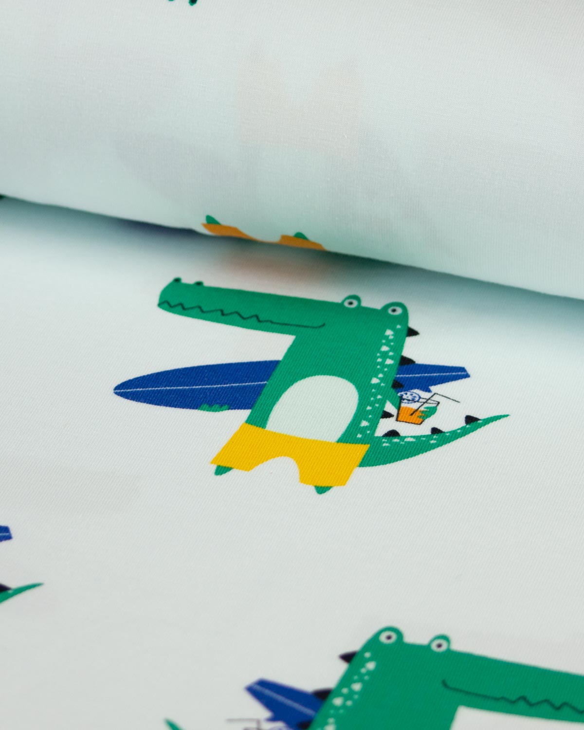 A close-up of a white cotton jersey  fabric printed with colorful cartoon crocodile motifs, including one wearing a yellow shirt and blue shorts while riding a blue surfboard.