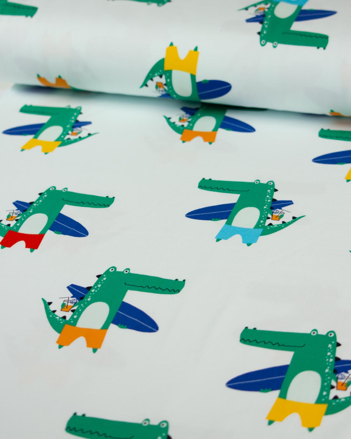 White cotton jersey  fabric with a repeating pattern of green alligators flying blue airplanes, with some alligators also featuring yellow, red, and light blue accents.