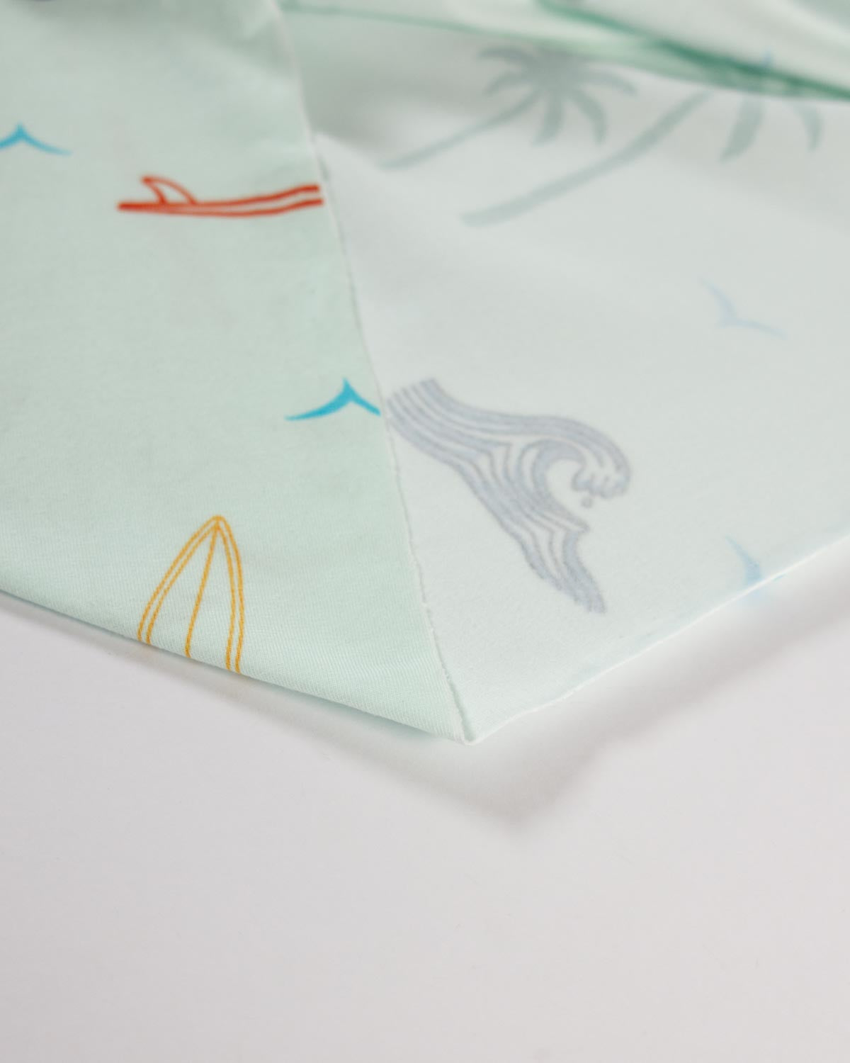 A close-up of a light mint green cotton jersey fabric featuring a subtle beach-themed print with palm trees, a gray wave, and small red, blue, and yellow accents.