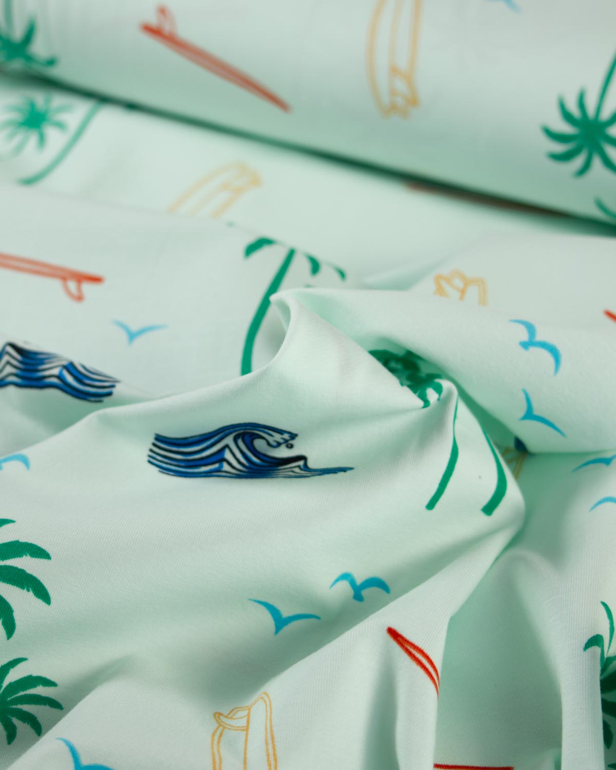 A close-up of light mint green cotton jersey fabric with a tropical beach print featuring blue waves, green palm trees, and small surfboards and birds.