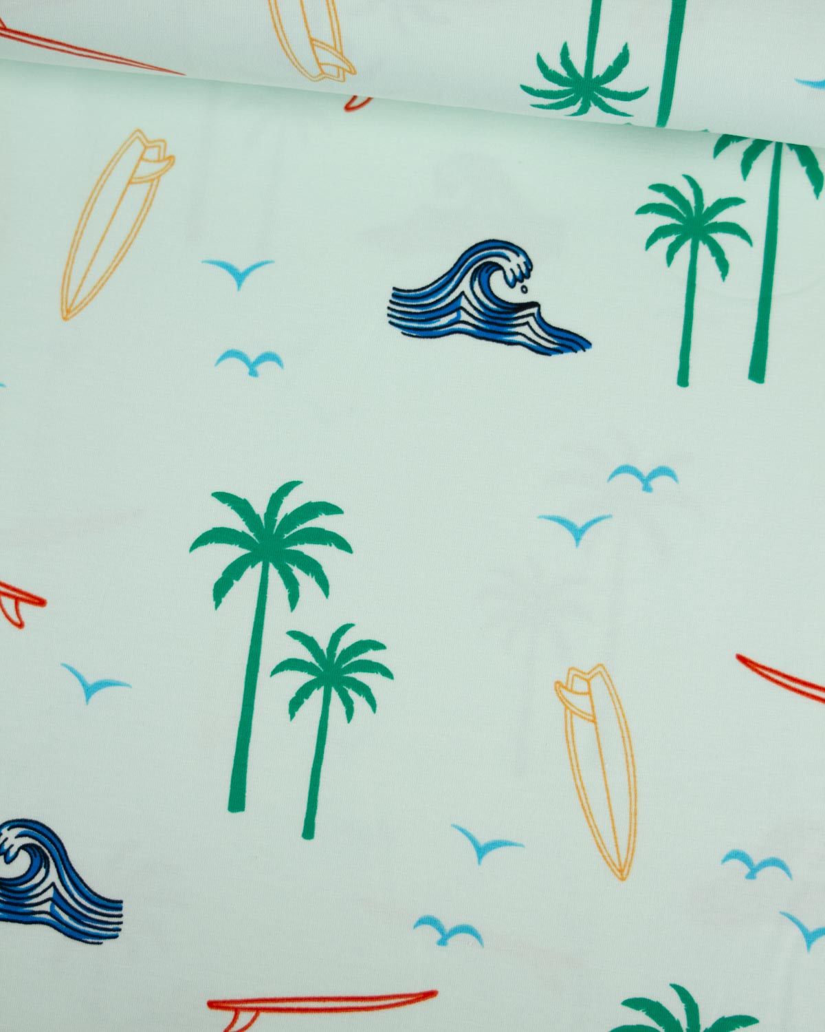 A close-up of a light mint green cotton jersey fabric with a tropical beach print featuring green palm trees, blue waves, yellow surfboards, and small blue birds.