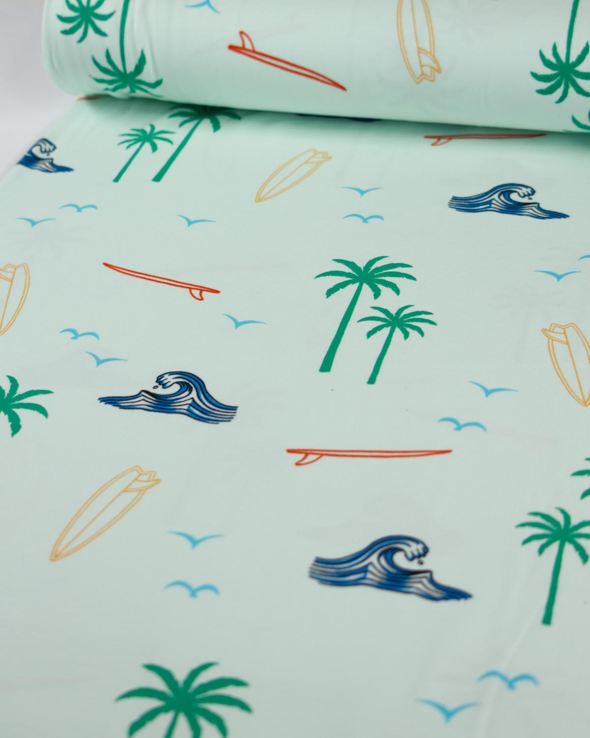 A roll of light mint green cotton jersey fabric with a tropical beach print, featuring palm trees, surfboards, waves, and birds, shown partially unrolled.