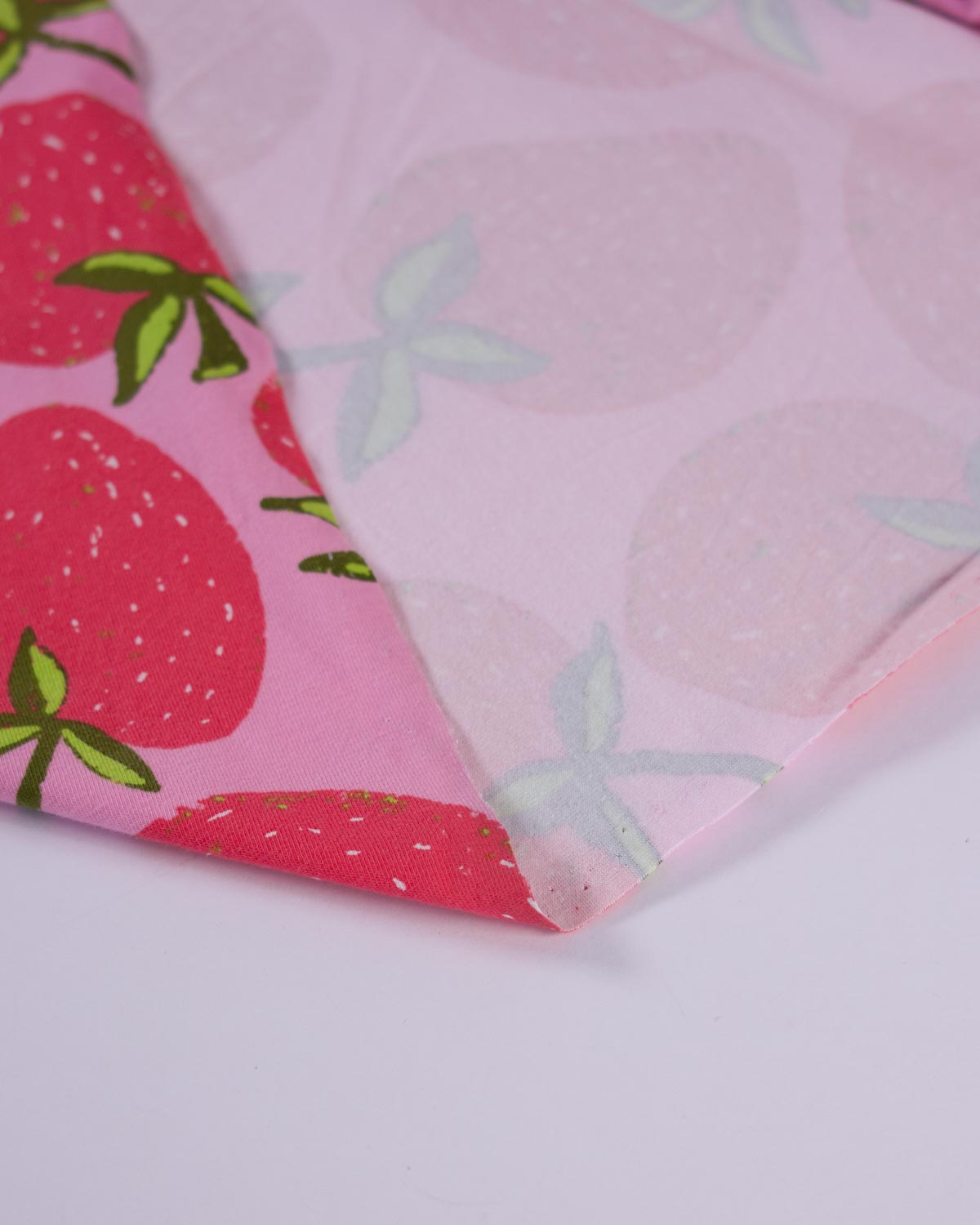 A close-up of a folded piece of pink patterned fabric, showing one side with a red strawberry print and the other side with a lighter pink strawberry pattern.