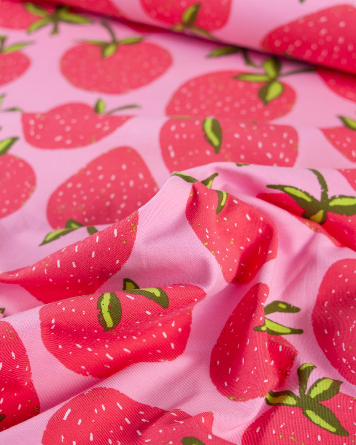 Pink cotton jersey fabric with a repeating strawberry print, shown in soft folds with close-up detail of the pattern.