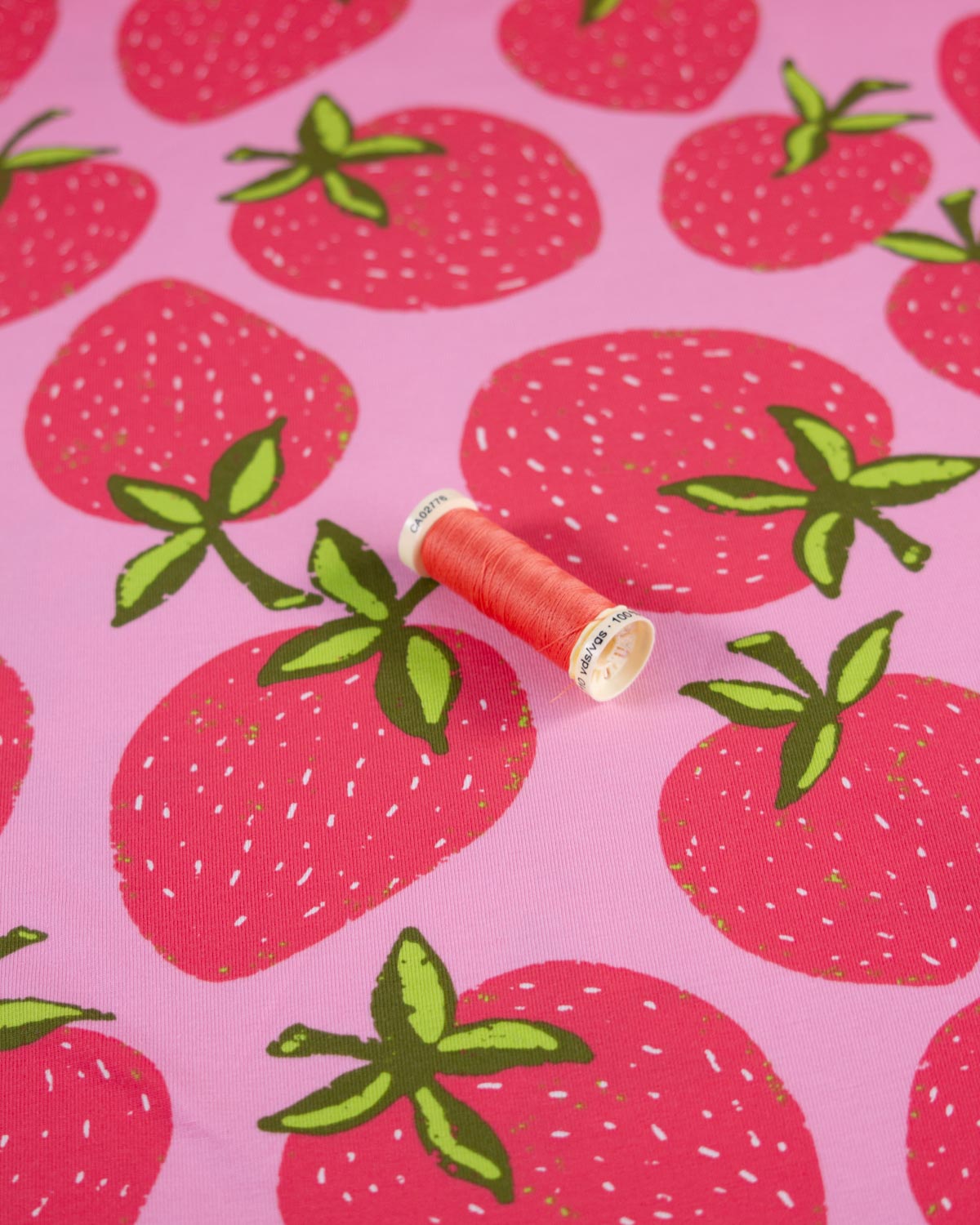 A close-up of a pink cotton jersey fabric surface printed with large red strawberries and green leafy tops, with a small spool of coral thread placed on top.