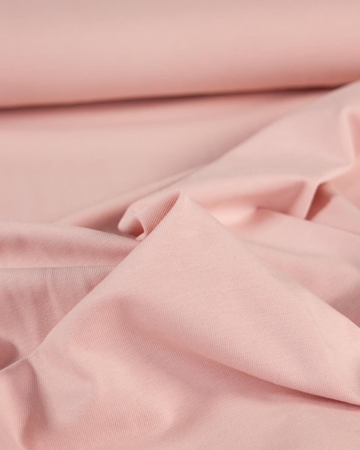 A close-up of soft blush pink organic cotton jersey fabric with gentle folds and a rolled section in the background.