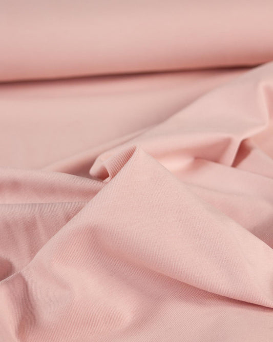 A close-up of soft blush pink organic cotton jersey fabric with gentle folds and a rolled section in the background.