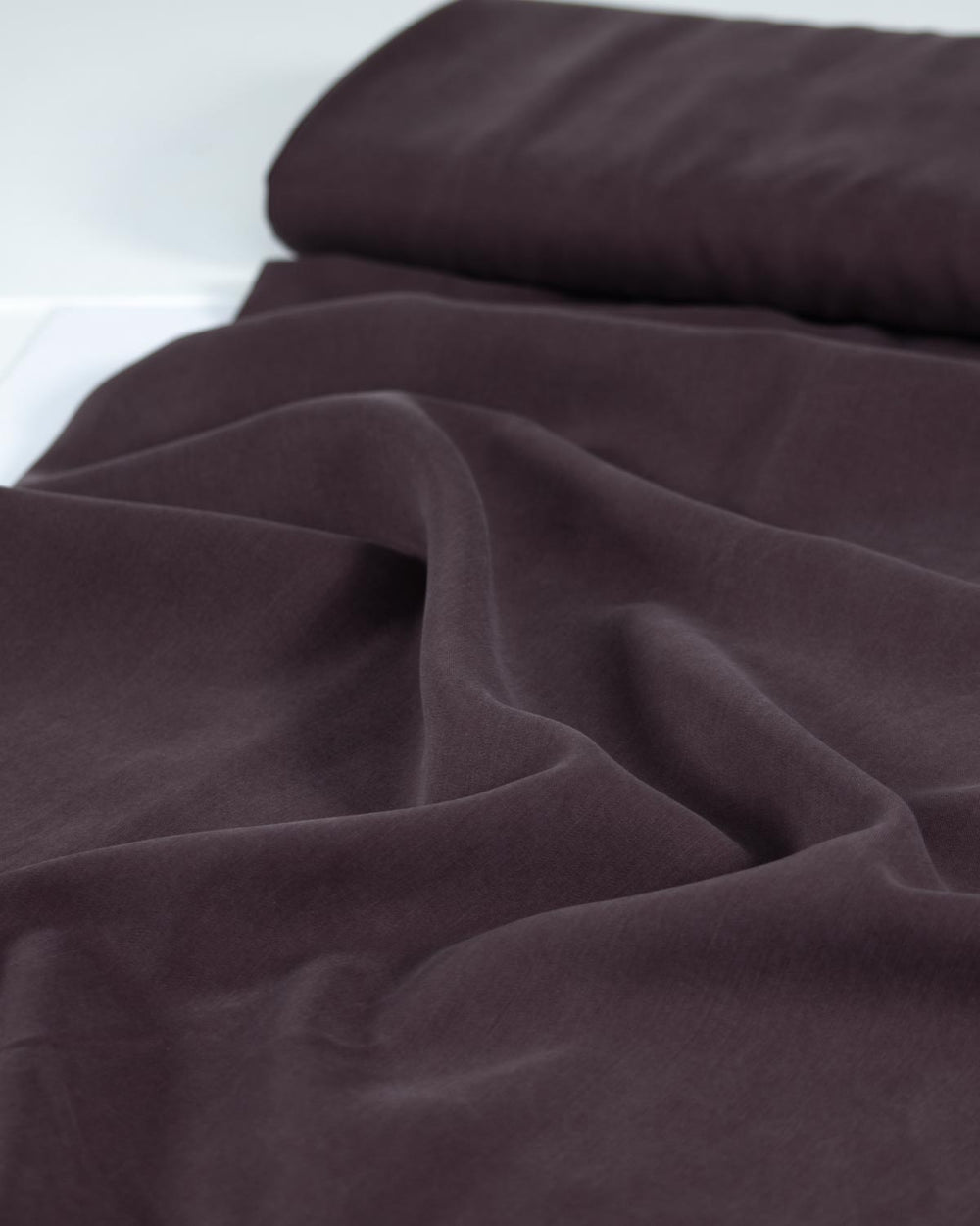 Dark brown draped tencel twill fabric with soft folds and a folded stack of the same material in the background.