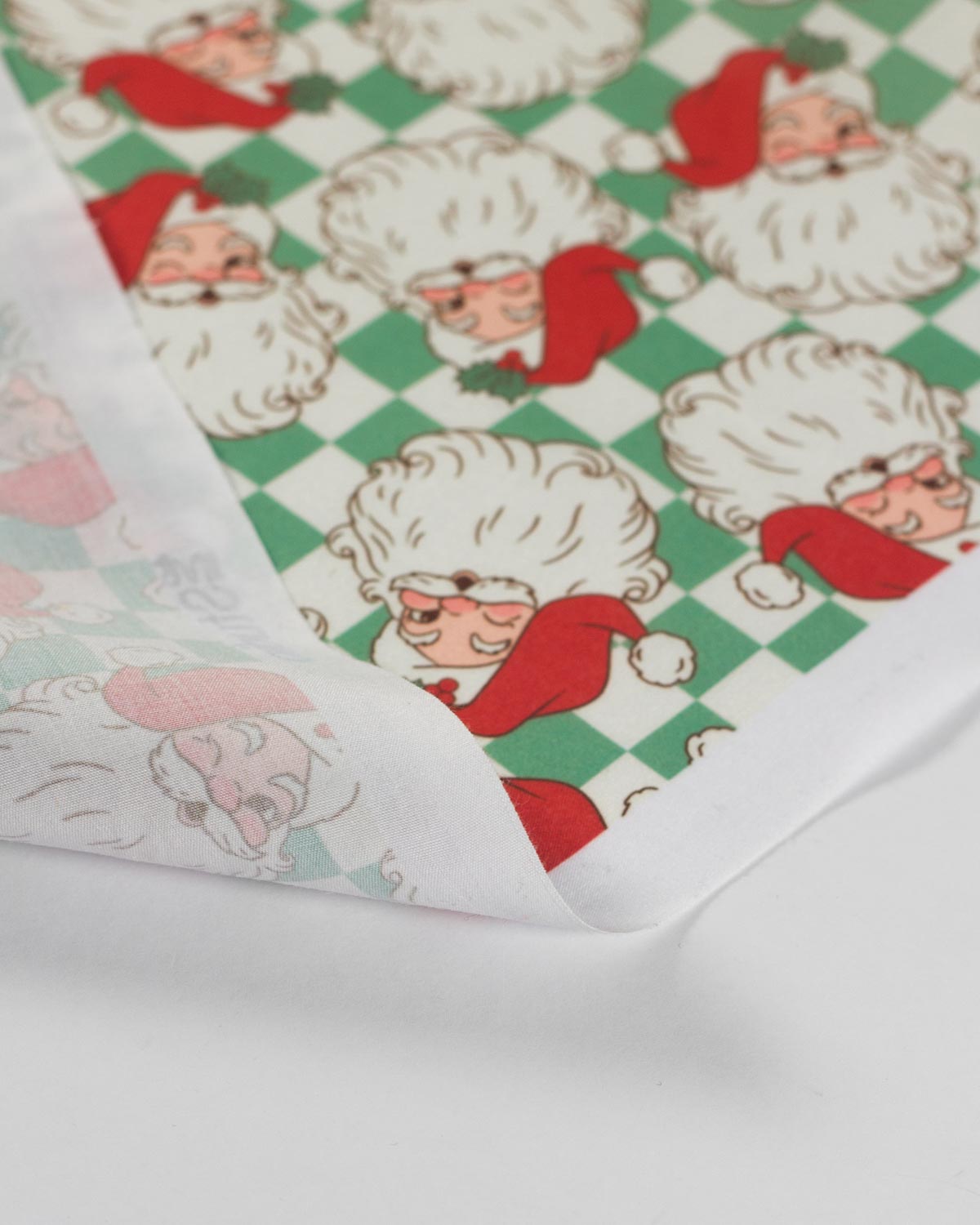 A close-up of a white fabric with a repeating Christmas Santa pattern, featuring a green checkered background and Santa faces in red hats and white hair, with one corner folded over to show the reverse side.