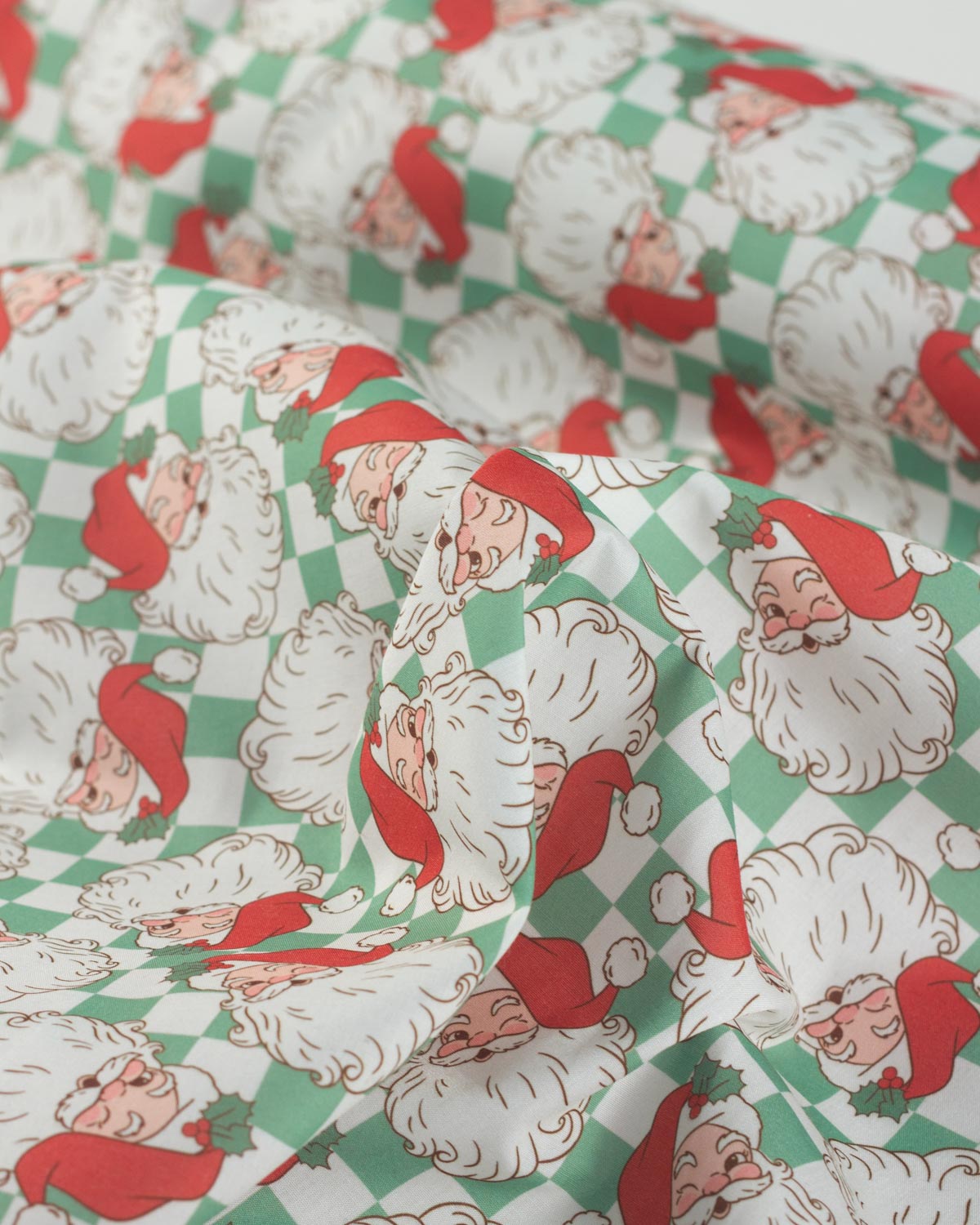 A close-up of a festive fabric featuring a repeating print of Santa Claus's face over a green and white checkered background.