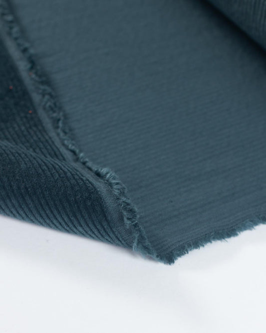 A close-up of a dark teal corduroy fabric folded over to show its texture and colour against a plain white background.