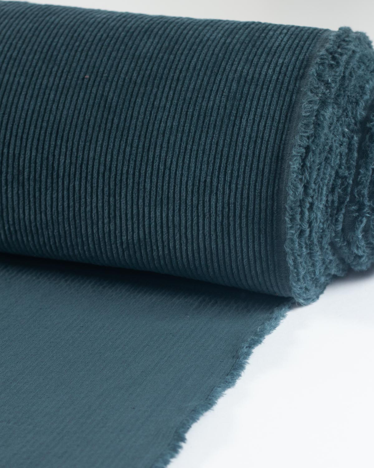 A close-up of a rolled piece of dark teal cotton corduroy fabric with a ribbed texture, with the unrolled section showing the selvedge on a white background.