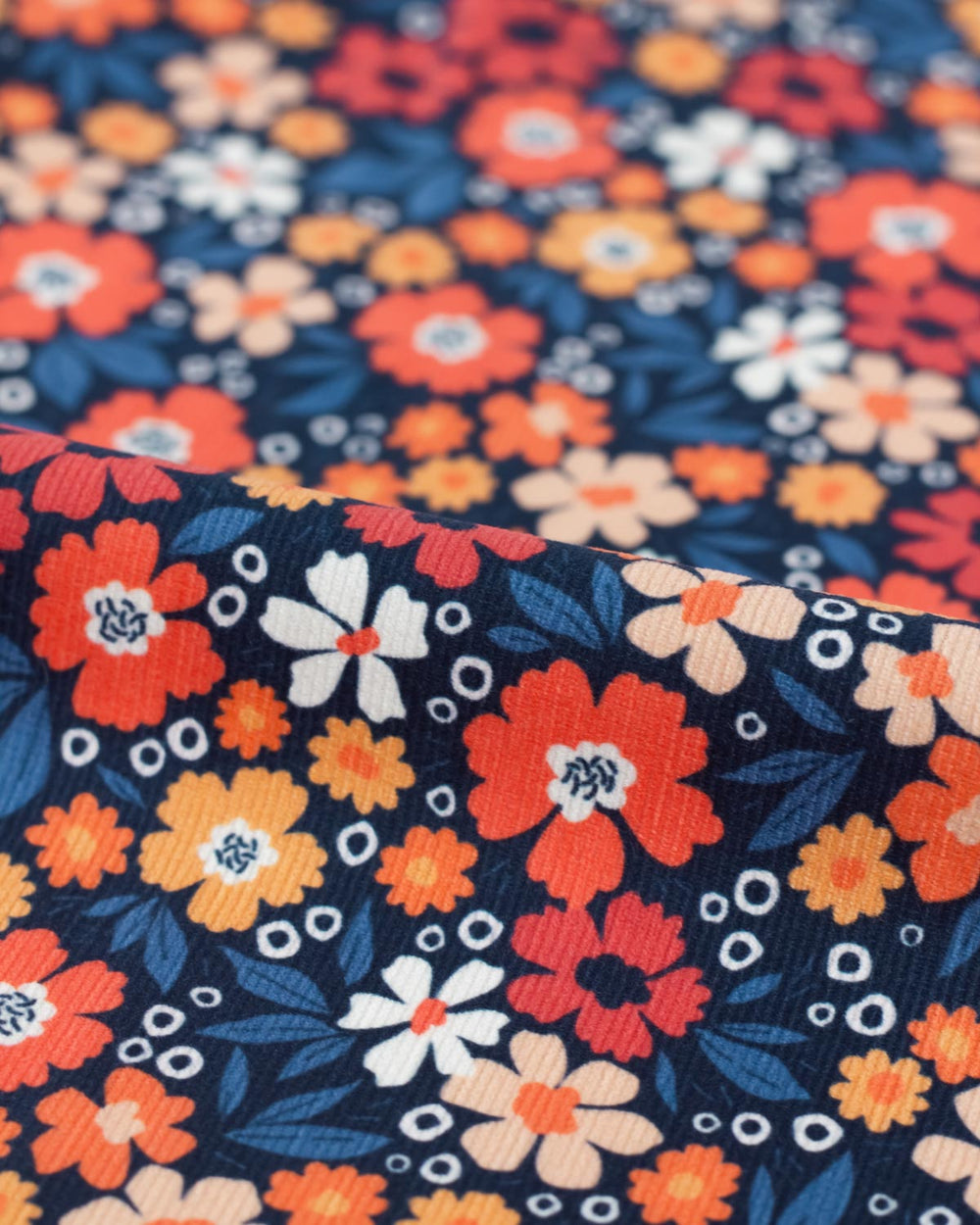 A close-up of a cotton babycord fabric with a dense floral pattern on a dark navy background. The design features orange, red, white, and peach flowers with blue leaves and small white circular accents.