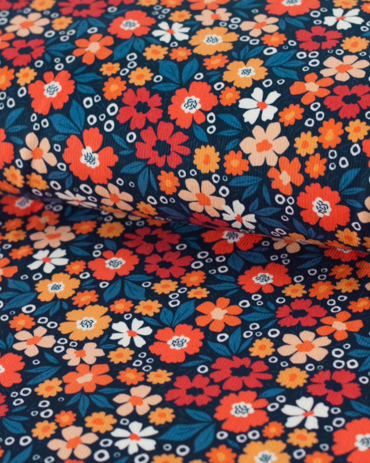 A close-up of a floral cotton babycord fabric with a dense pattern of orange, red, white, and yellow flowers with teal leaves on a dark navy background.