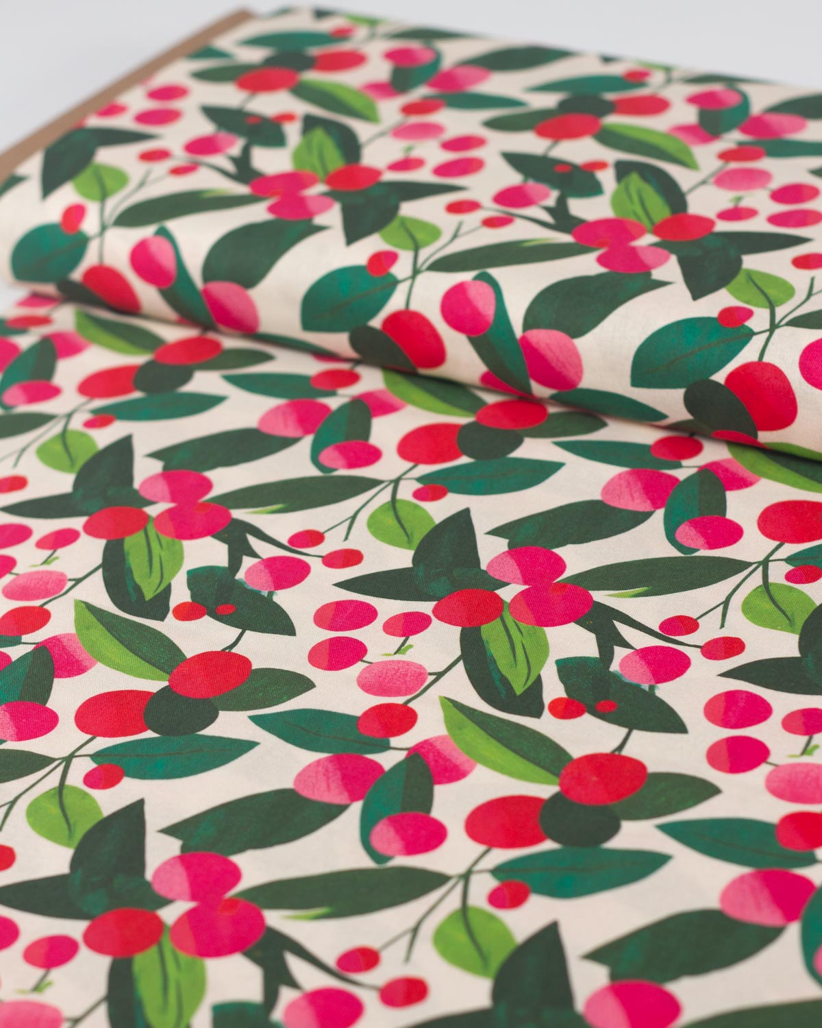 A close-up of a christmas cotton fabric featuring a repeating botanical pattern of red berries, pink berries, and green leaves on a warm cream background.