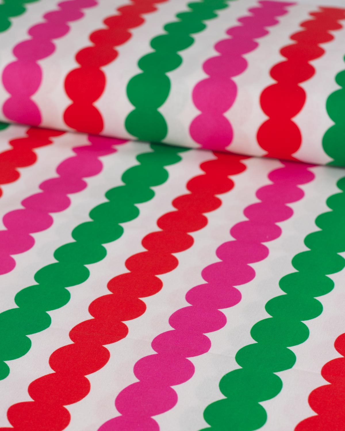 A close-up of a white cotton fabric  with repeating diagonal rows of scalloped red, green, and pink shapes.