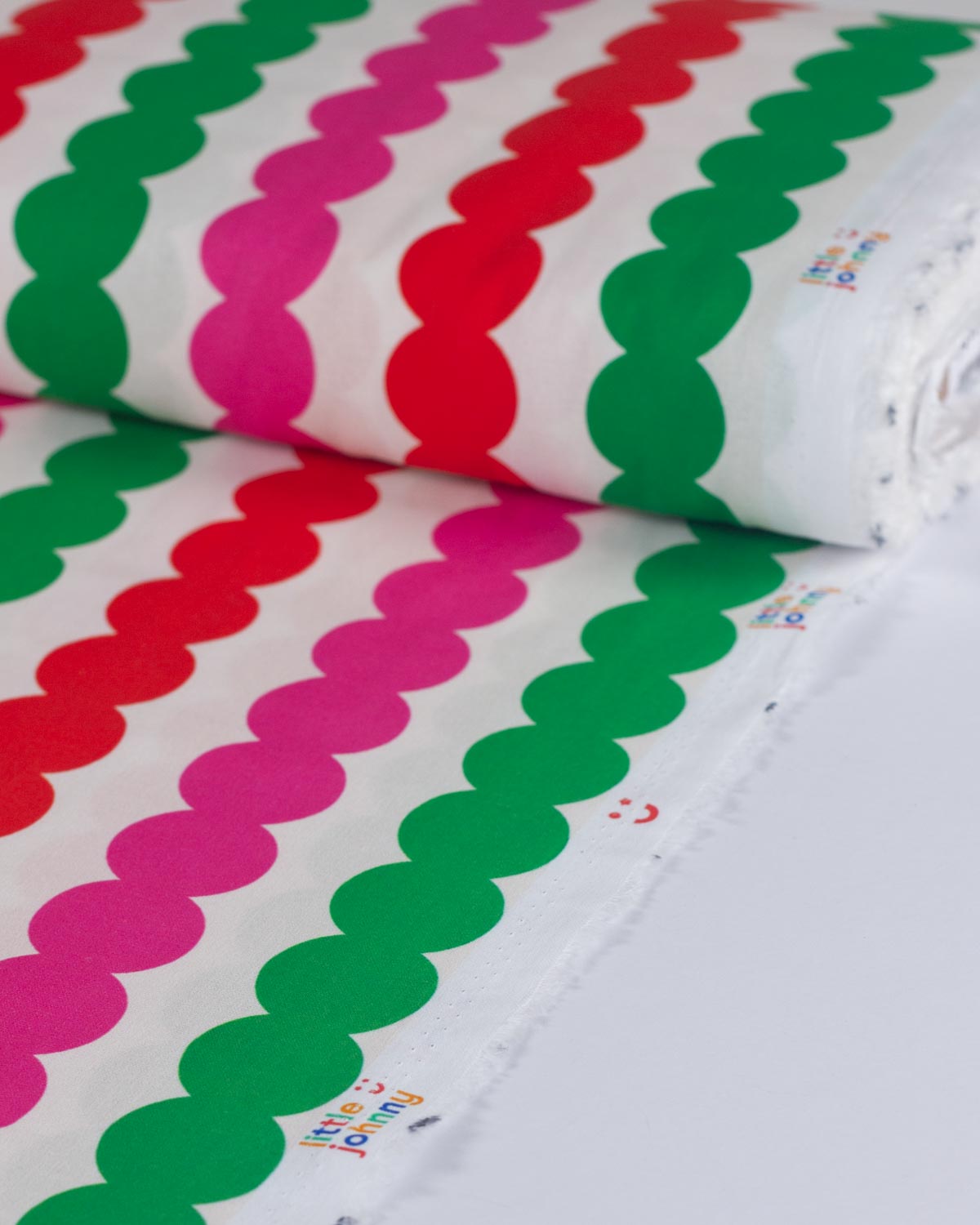 A close-up of a white fabric roll featuring repeating diagonal stripes of scalloped green and pink shapes, with small colorful printed text near the lower edge.