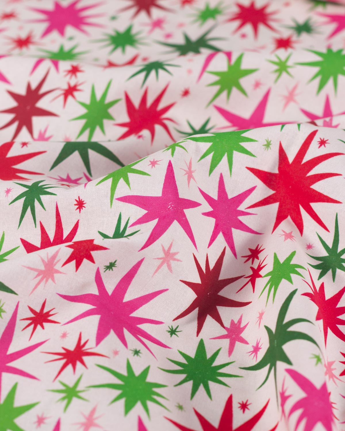A close-up of patterned fabric with large and small starburst shapes in bright pink, red, and green on a light off-white background.