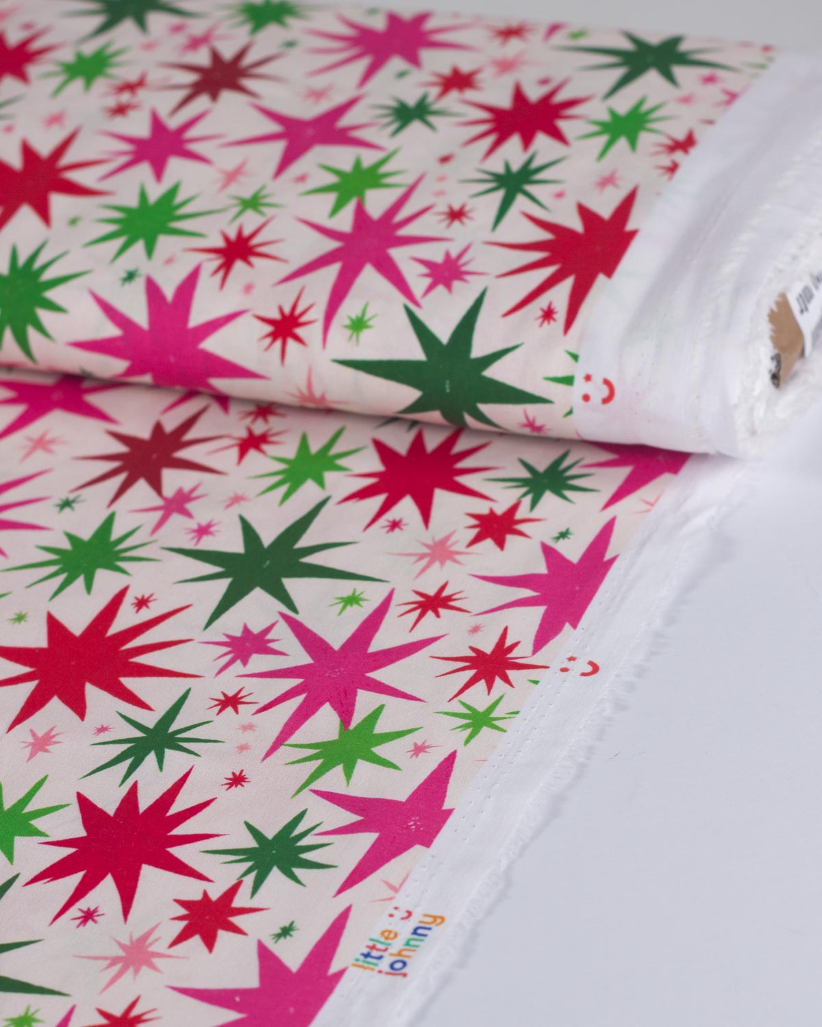 A roll of white fabric printed with a festive pattern of red, pink, and green starburst shapes, shown on a white surface.