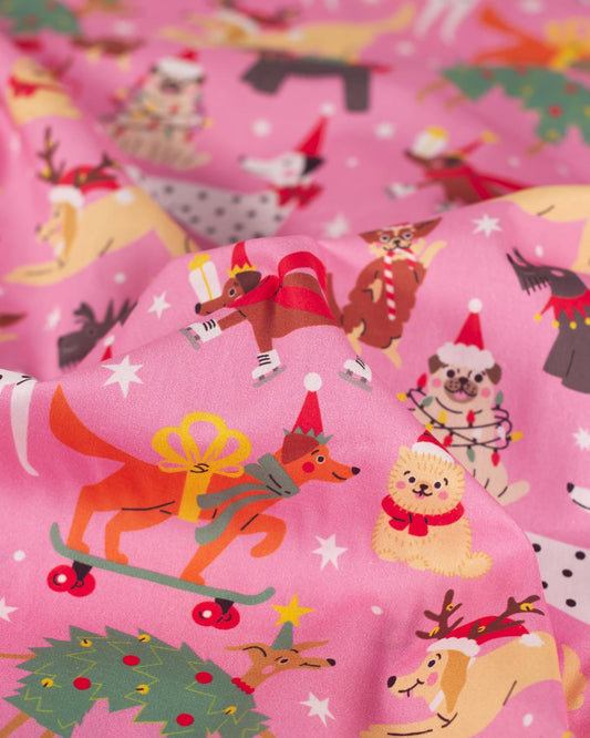 Pink fabric with a festive Christmas print featuring illustrated dogs, Santa hats, stars, and holiday greenery in warm multicolor tones.