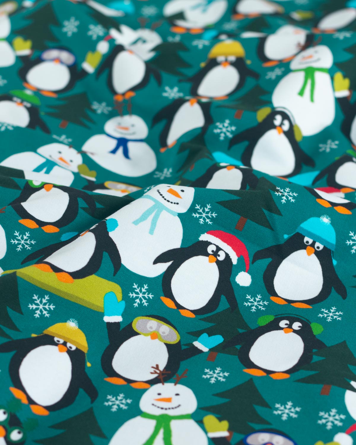 A close-up of festive holiday jersey fabric featuring a repeating pattern of penguins, snowmen, snow trees, and snowflakes on a teal background.